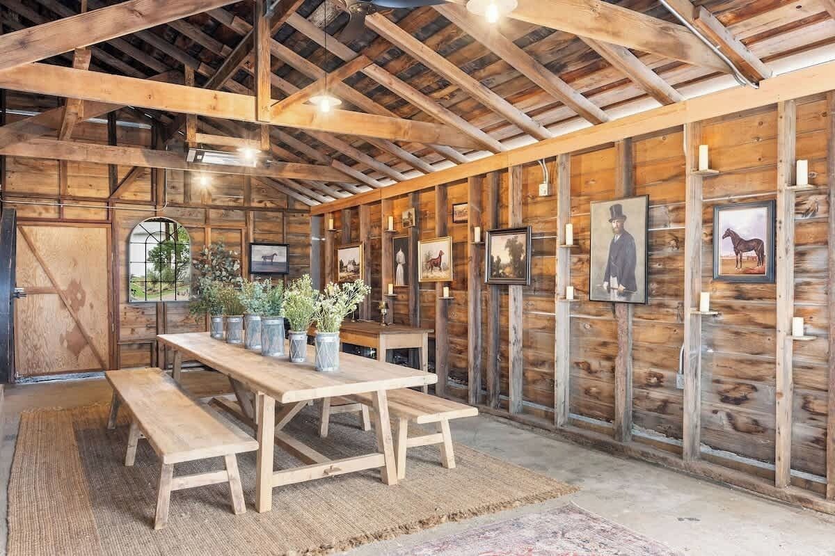A rustic barn-style interior with wooden beams, a long table with benches, and framed pictures on the walls.