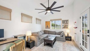 A modern living room with a sectional sofa, a wooden desk, a ceiling fan, and a large window. - The Cypress Bungalows (Sonoma)