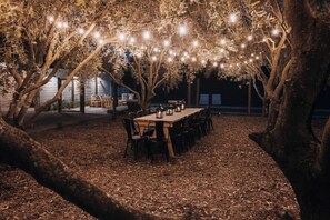 An outdoor dining area with a long table and chairs, surrounded by trees and string lights.
