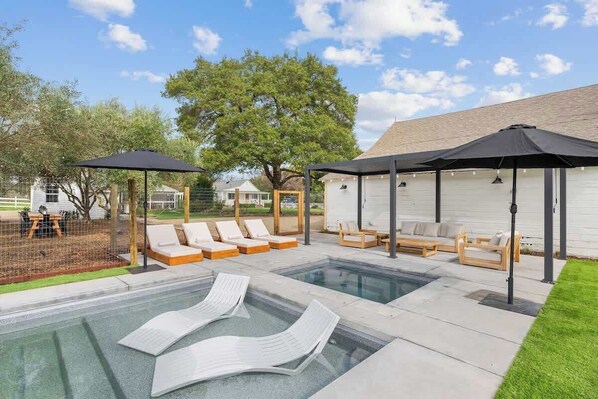 A swimming pool with lounge chairs and umbrellas, surrounded by a fenced area with a building in the background.
