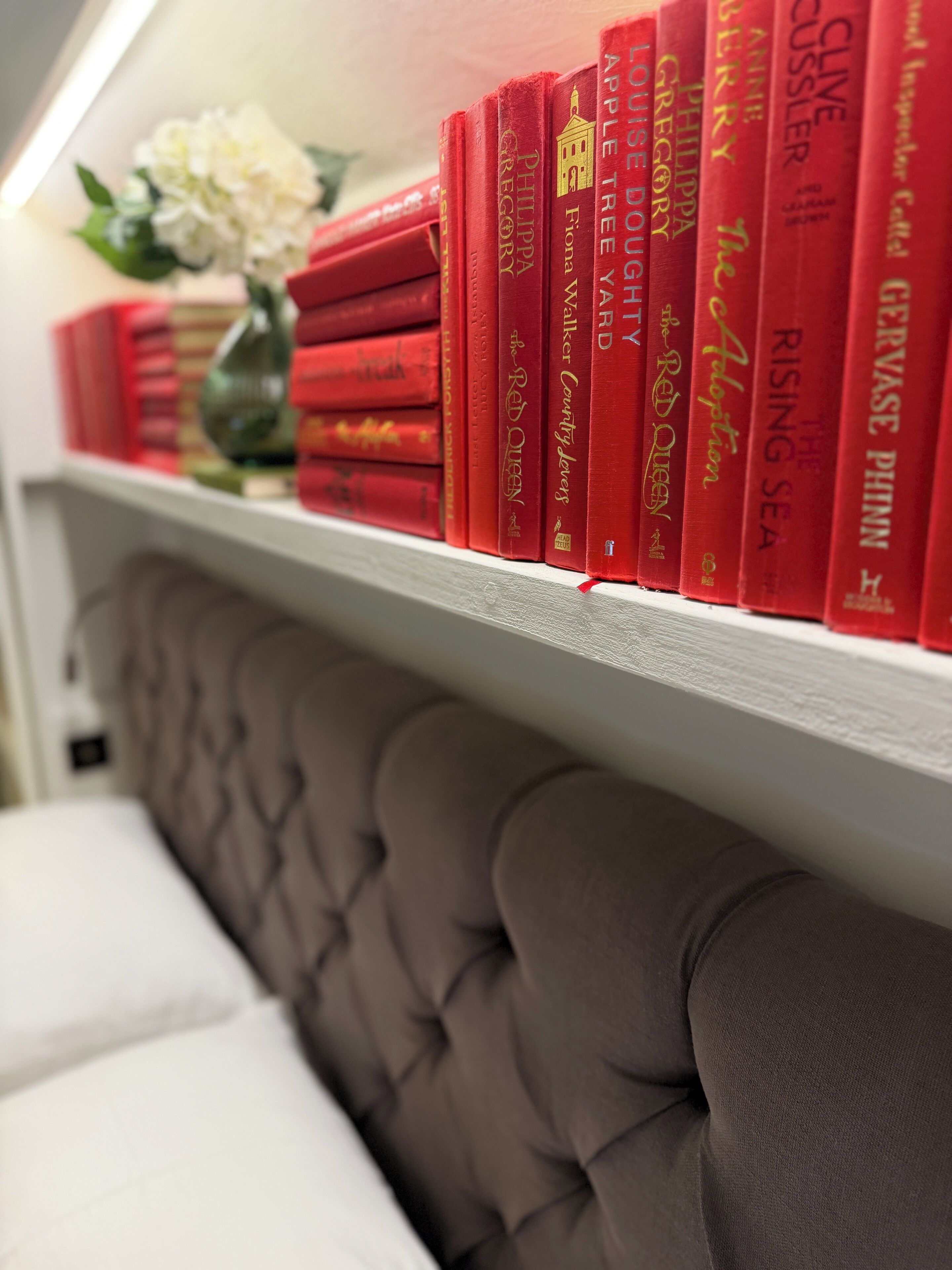 Una estantería con libros rojos y un florero con flores blancas.