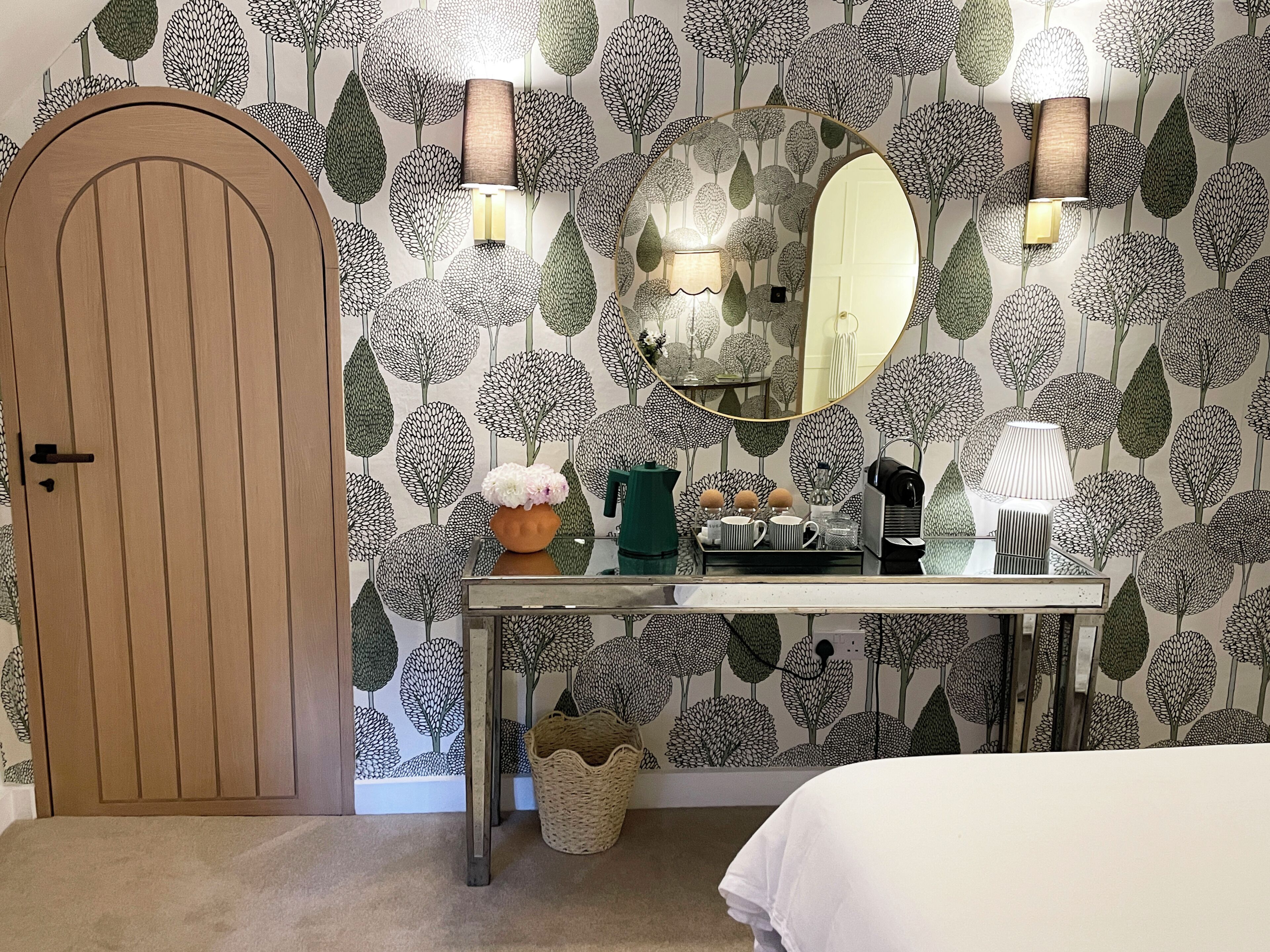 Una habitación con puerta de madera, un espejo redondo, una mesa con varios objetos y una pared con diseño de hojas.