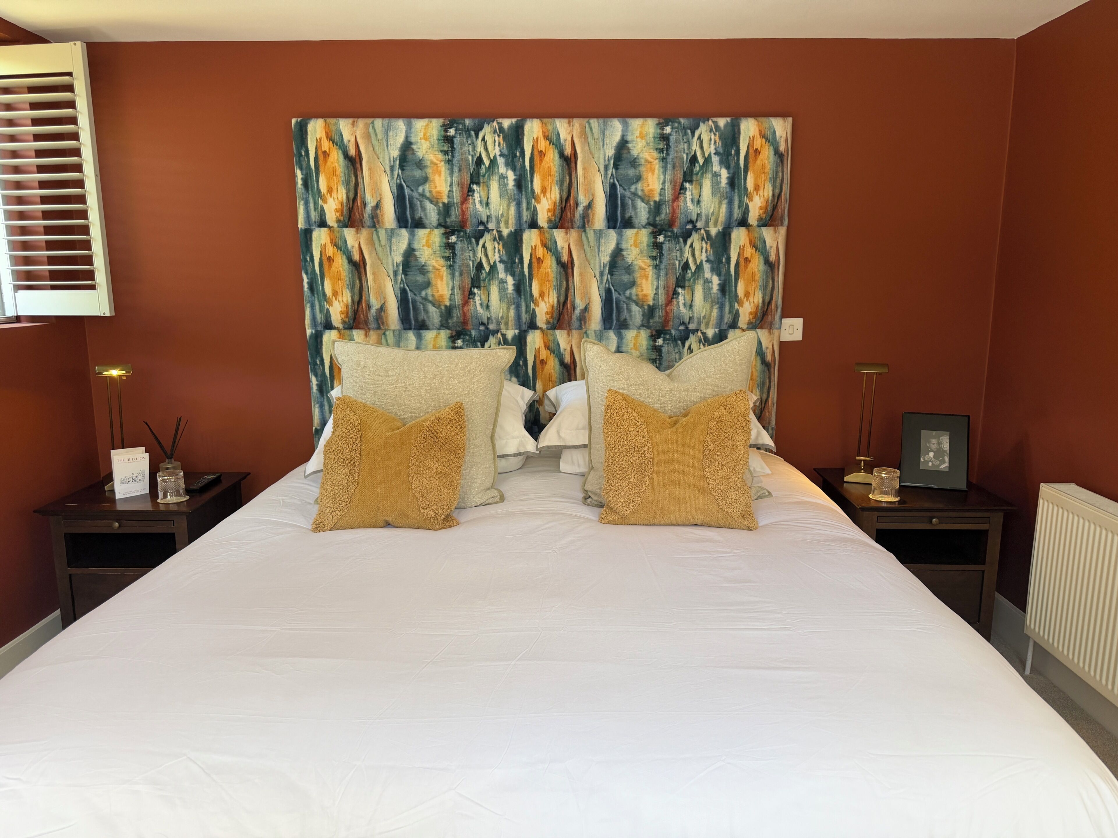 A hotel room with a large bed, two nightstands, a headboard with a colorful abstract design, and a window with blinds.