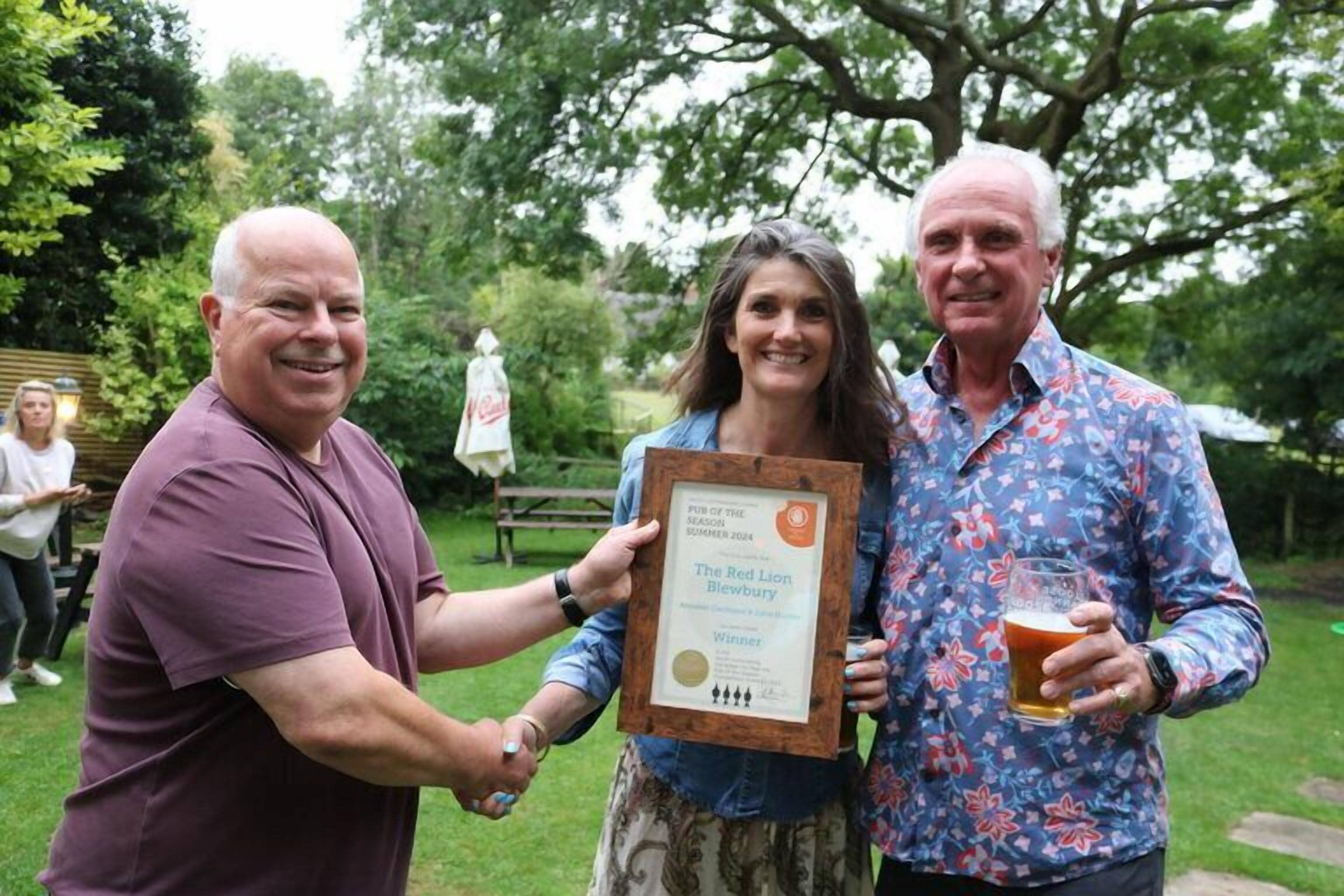 Three people holding a framed award in an outdoor setting with trees and a picnic table in the background.