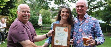 Three people holding a framed award in an outdoor setting with trees and a picnic table in the background.
