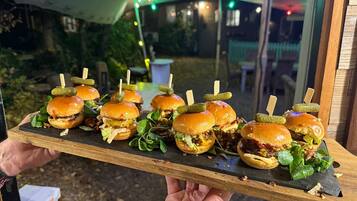 Una bandeja con mini hamburguesas adornadas con pepinillos y vegetales verdes, sostenida por una persona.