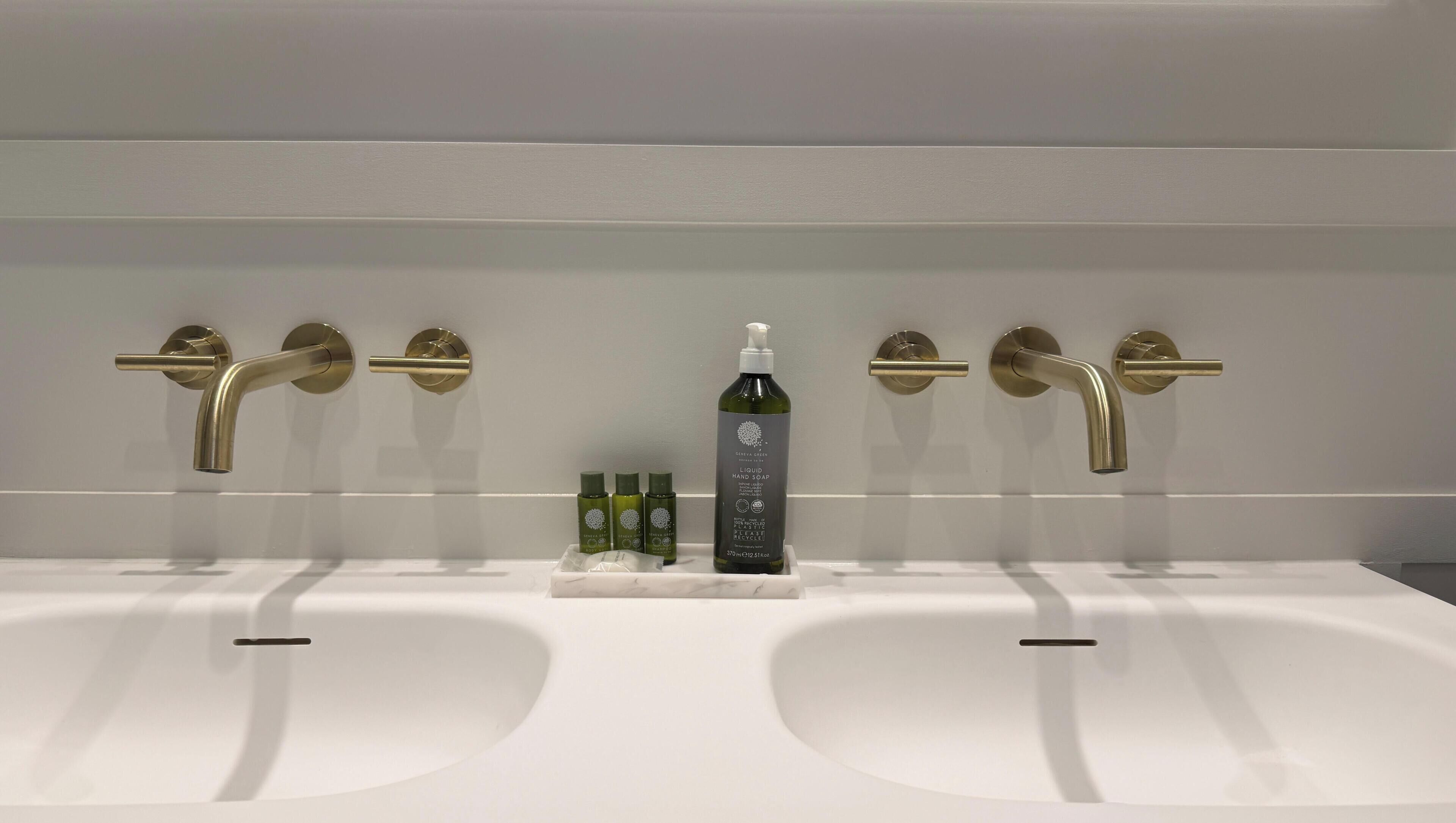 A bathroom with two sinks, a bottle of hand soap, and three green bottles on the countertop.