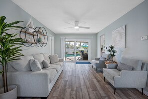 Uma sala de estar moderna com um sofá cinzento, duas poltronas, um ventilador de teto e piso de madeira. Há uma janela grande com vista para uma piscina e móveis de exterior.