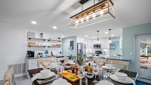 Une cuisine moderne avec des meubles blancs, un coin repas doté d’une table en bois pour quatre personnes, et une vue sur le salon attenant.