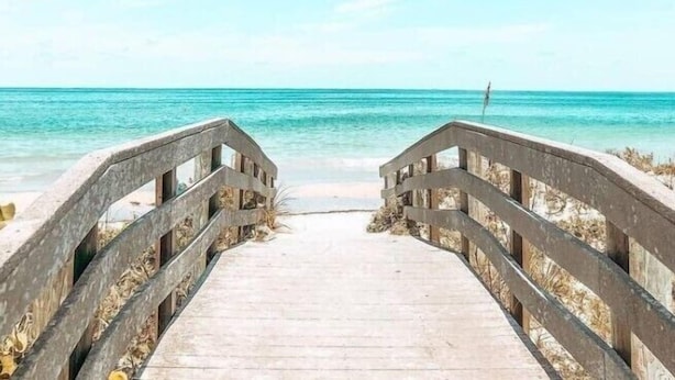 Une promenade en bois mène à une plage où l’eau est d’un bleu turquoise limpide et le ciel est dégagé.