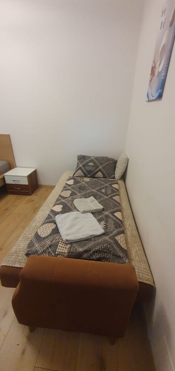 Una habitación pequeña con cama individual, colcha estampada, un banco marrón y una mesita de noche.