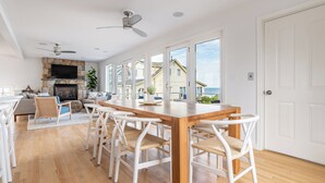 Un vaste espace de vie comprenant une table à manger en bois, des chaises blanches, une cheminée et de grandes fenêtres offrant une vue sur la mer.