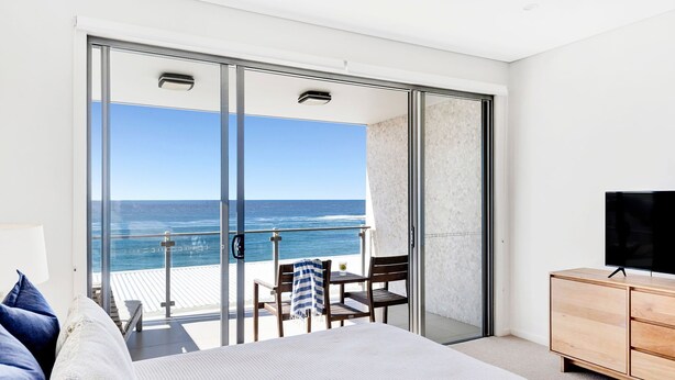 A balcony with a glass door leading to a room with a bed, a TV, and a wooden cabinet. - White Sails 3 - Oceanfront Luxury, Pool & Convent Beach Views - Holiday Yamba (Yamba)