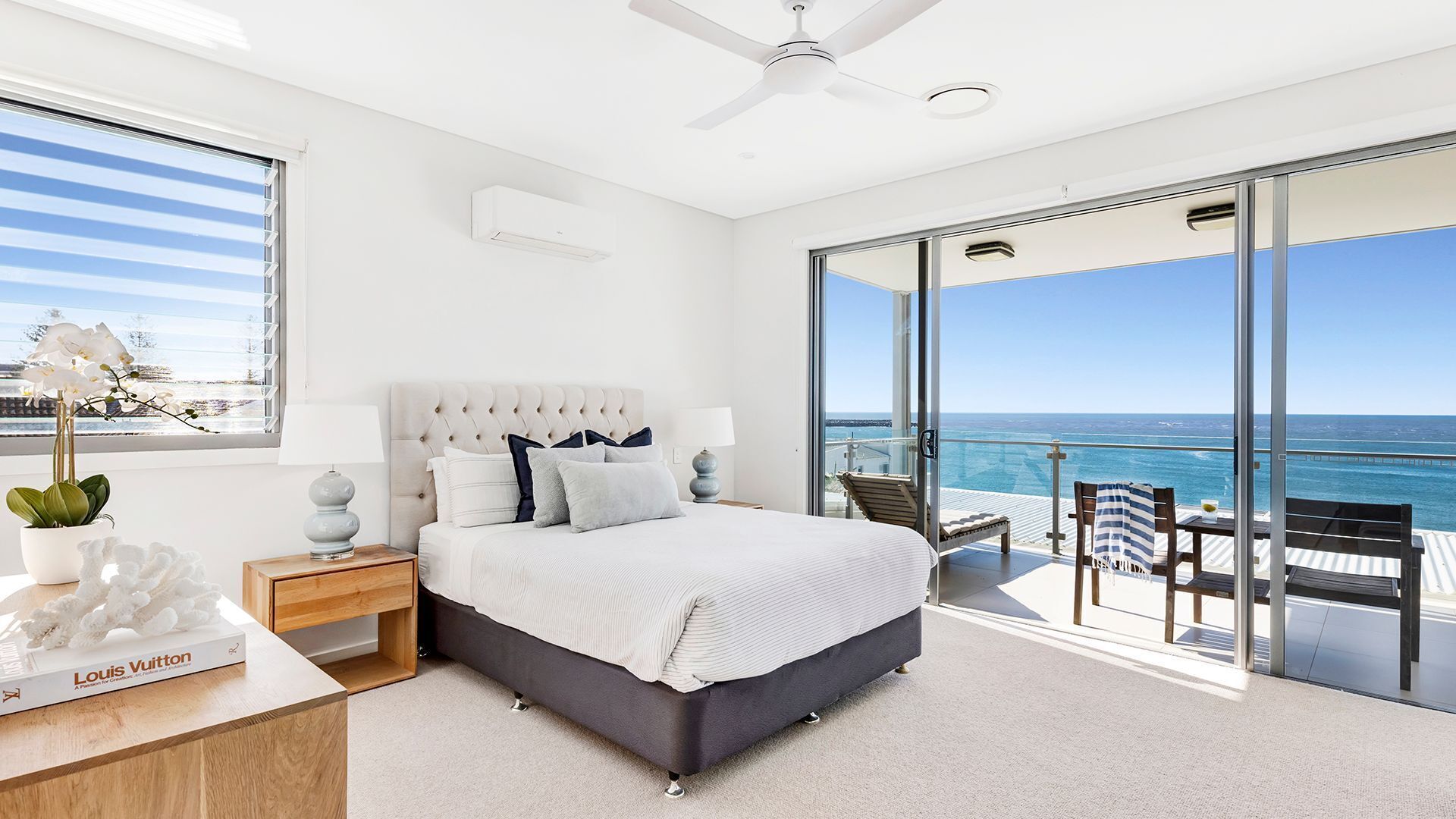 A bedroom with a large bed, a nightstand, a lamp, a Louis Vuitton book, and a view of the ocean through a sliding glass door.