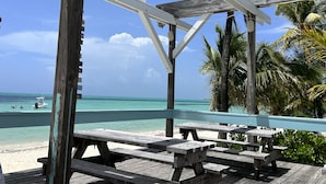 Une terrasse en bois avec des tables de pique-nique, donnant sur une plage aux eaux turquoise et cristallines, bordée de palmiers.