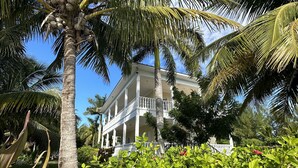 Un grand bâtiment de style colonial, peint en blanc, doté d’un balcon et entouré d’une végétation luxuriante et de palmiers.