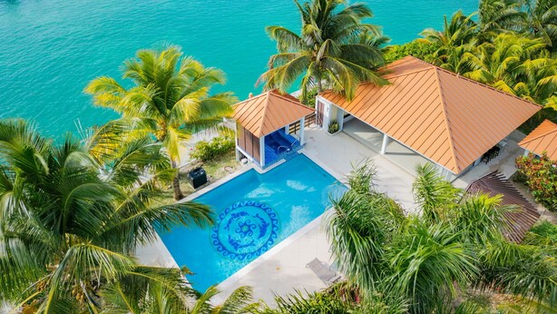 Vue aérienne d’un complexe hôtelier doté d’une piscine entourée de palmiers, avec en arrière-plan une mer turquoise.
