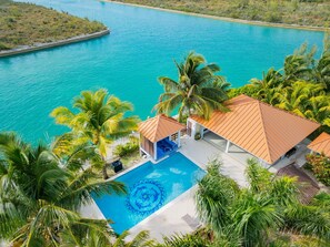 Luchtbeeld van een resort met een zwembad, omgeven door palmbomen, met uitzicht op een turquoise zee.