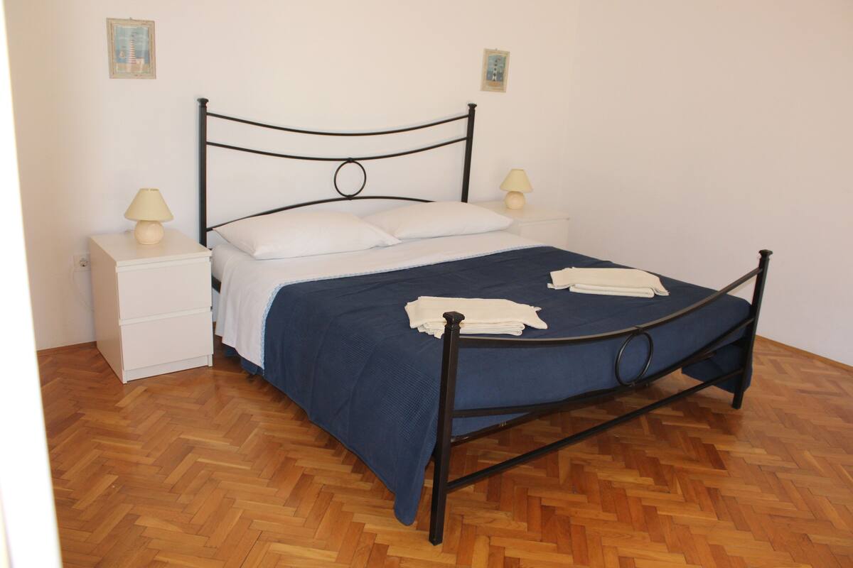 Cama com estrutura de metal, roupa de cama branca e manta azul. Há duas mesas de cabeceira com abajures.