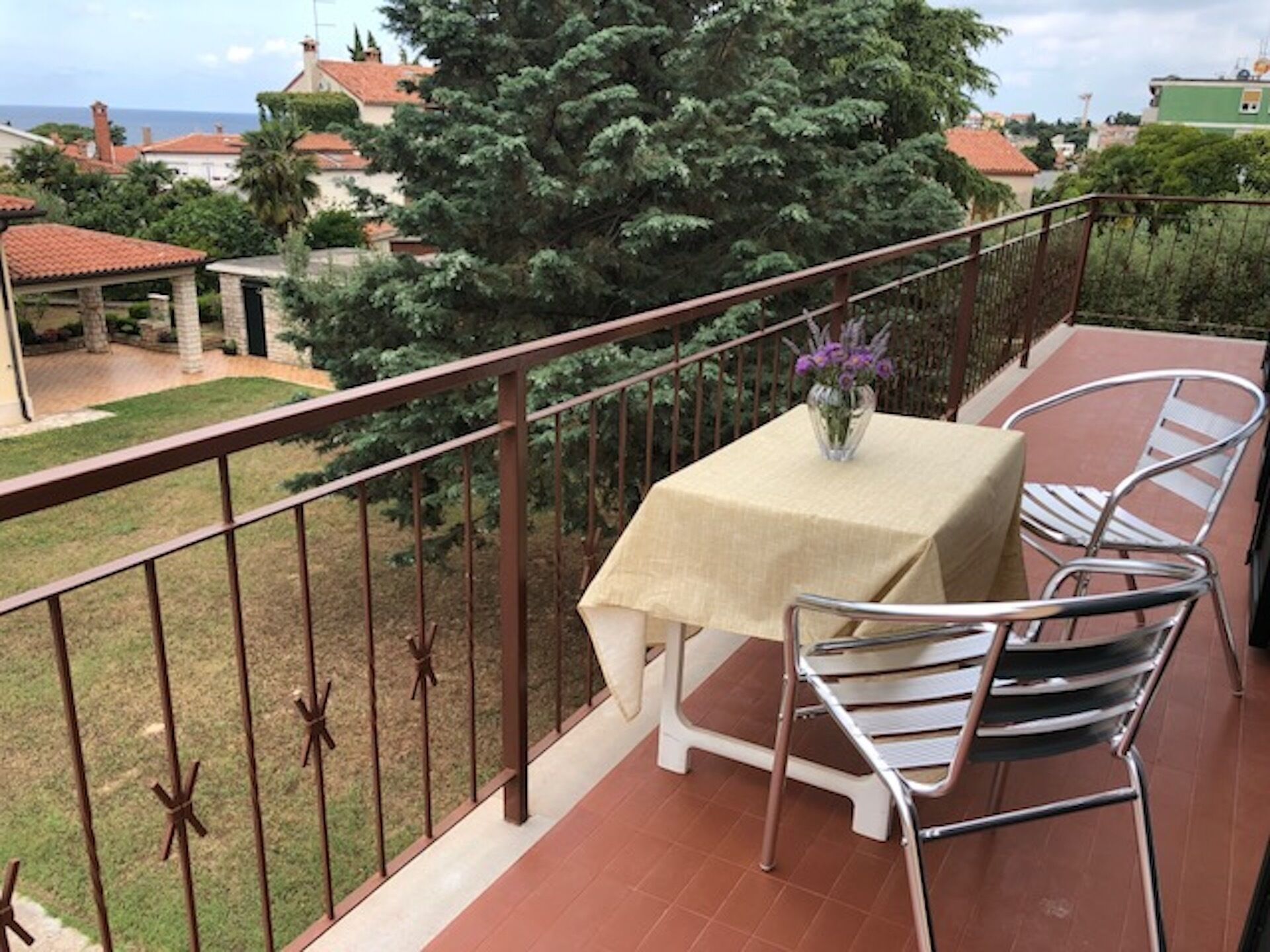 Un balcón con mesa y sillas, barandilla, y vistas a una zona residencial con árboles y casas.
