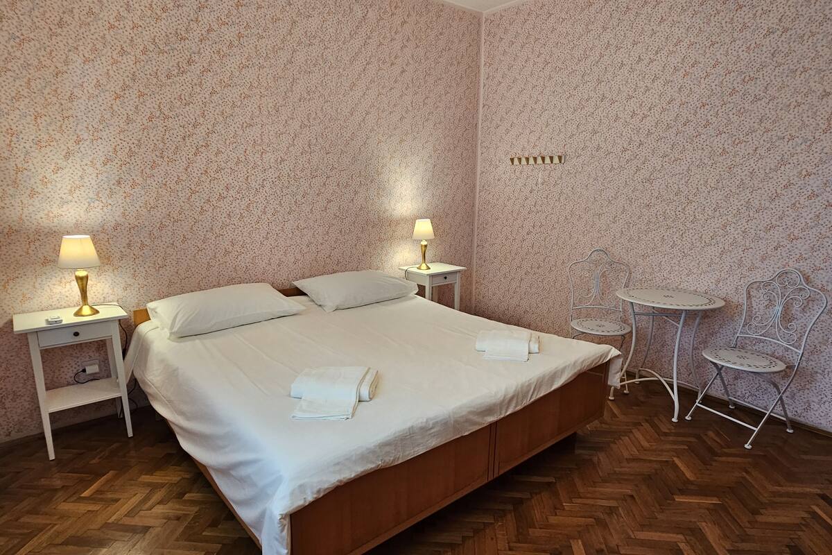 Quarto com cama de madeira, duas mesas de cabeceira com abajures, uma mesinha redonda e duas cadeiras.