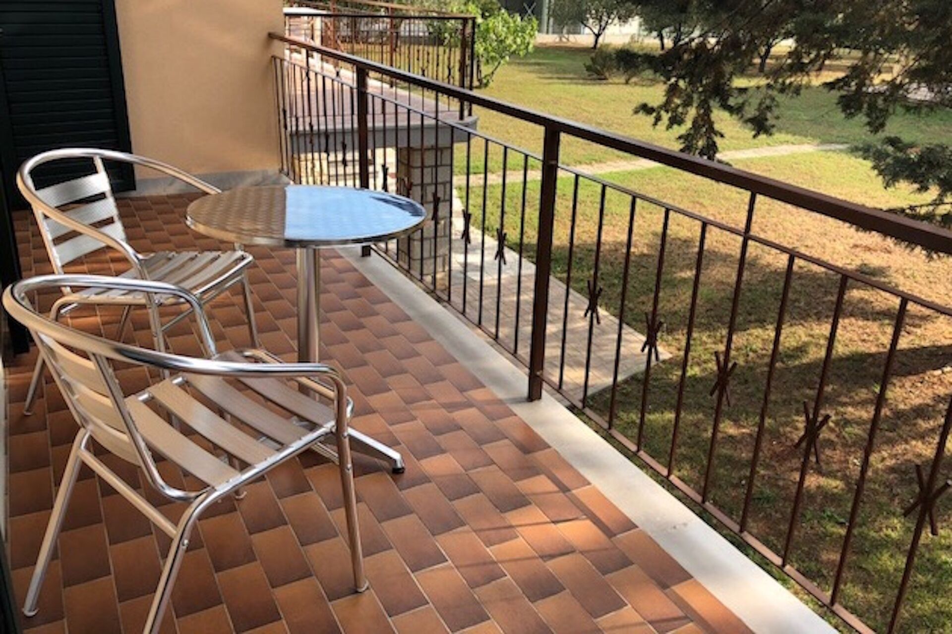 A balcony with two metal chairs and a small table, a tiled floor, and a metal railing overlooking a grassy area.