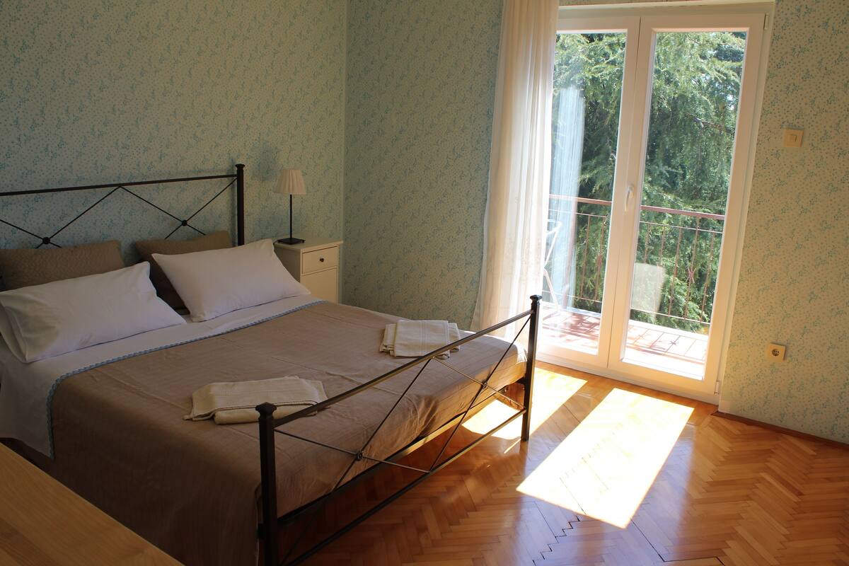 Quarto com cama, piso de madeira e vista através de uma porta de vidro.