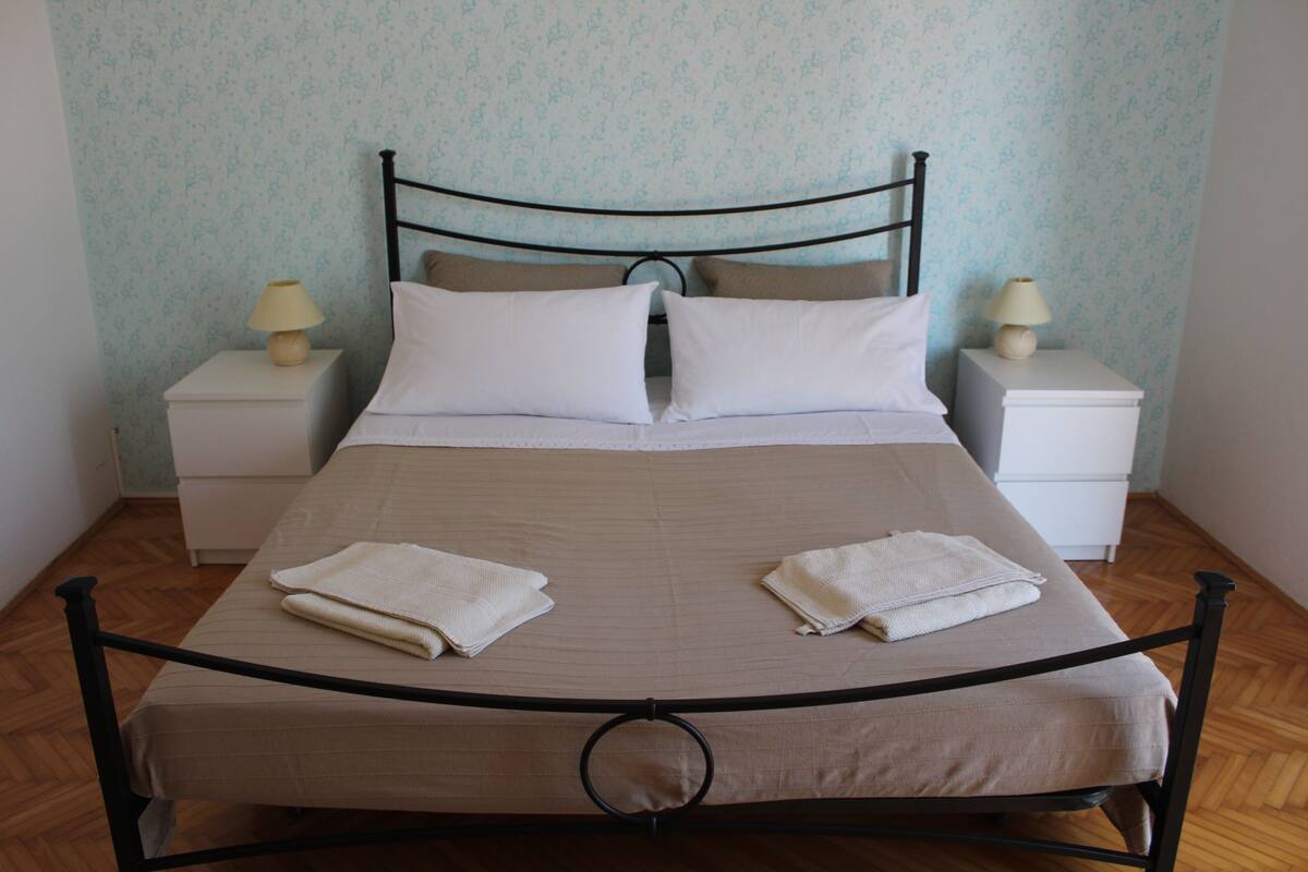 Cama bem arrumada com estrutura de metal, duas almofadas e duas toalhas dobradas. Há duas mesinhas de cabeceira com luminárias de cada lado da cama.