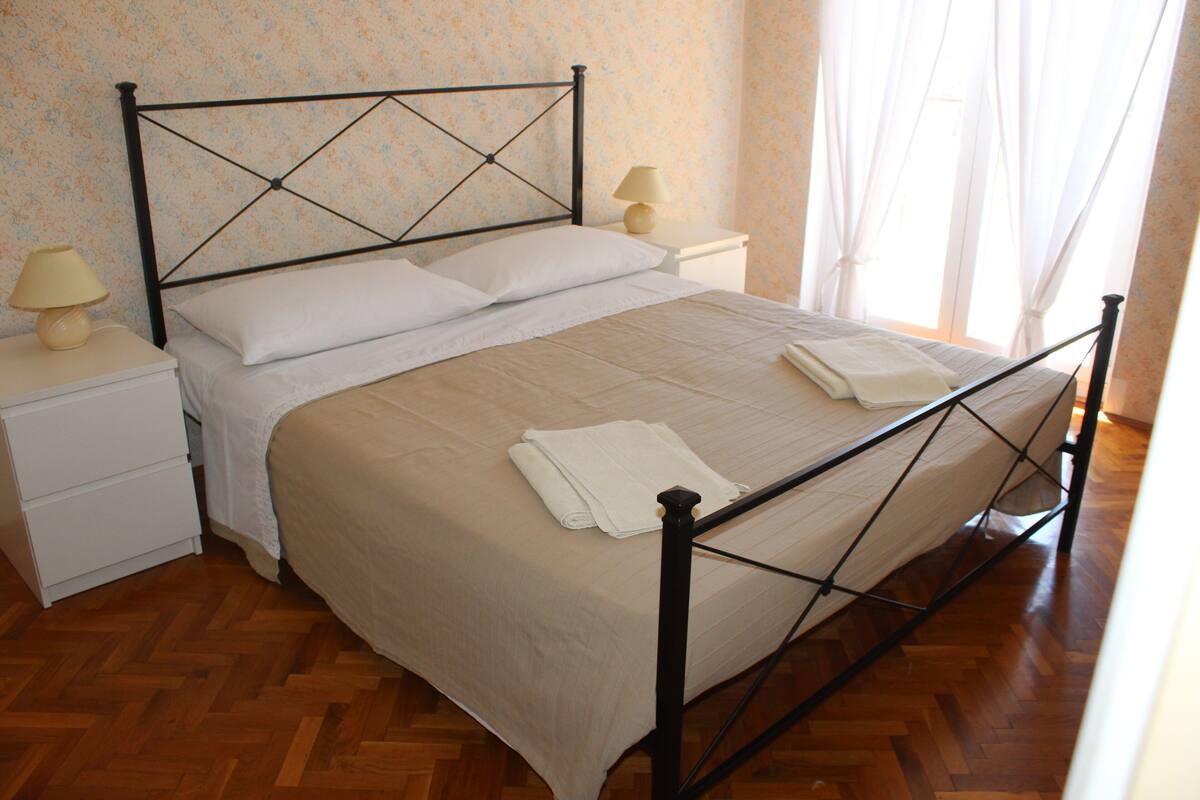 Uma cama bem arrumada com estrutura de metal, uma mesa de cabeceira com abajur e piso de madeira.