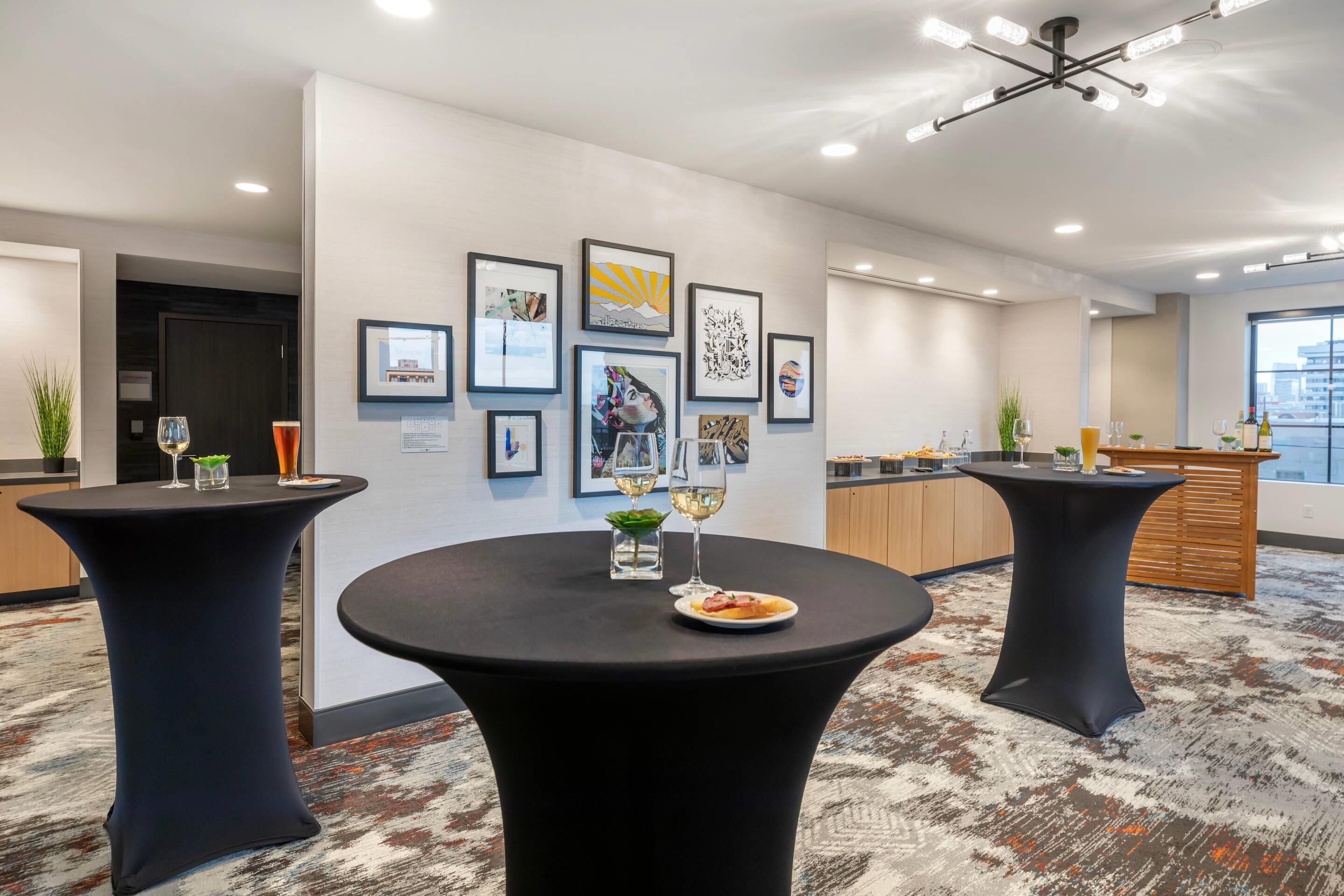 A modern hotel lobby with round tables, framed artwork on the walls, and a view of the city through large windows.