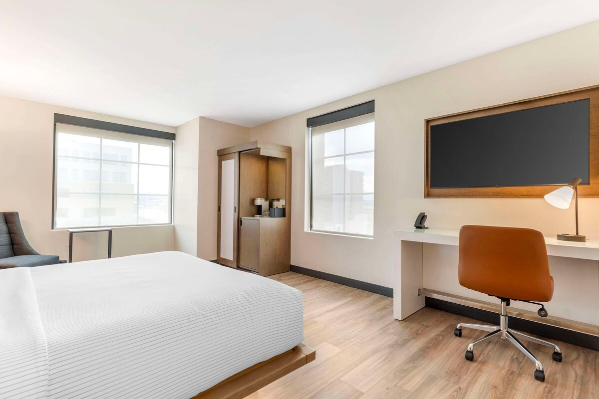 Quarto de hotel com cama, escrivaninha, cadeira e uma janela grande.