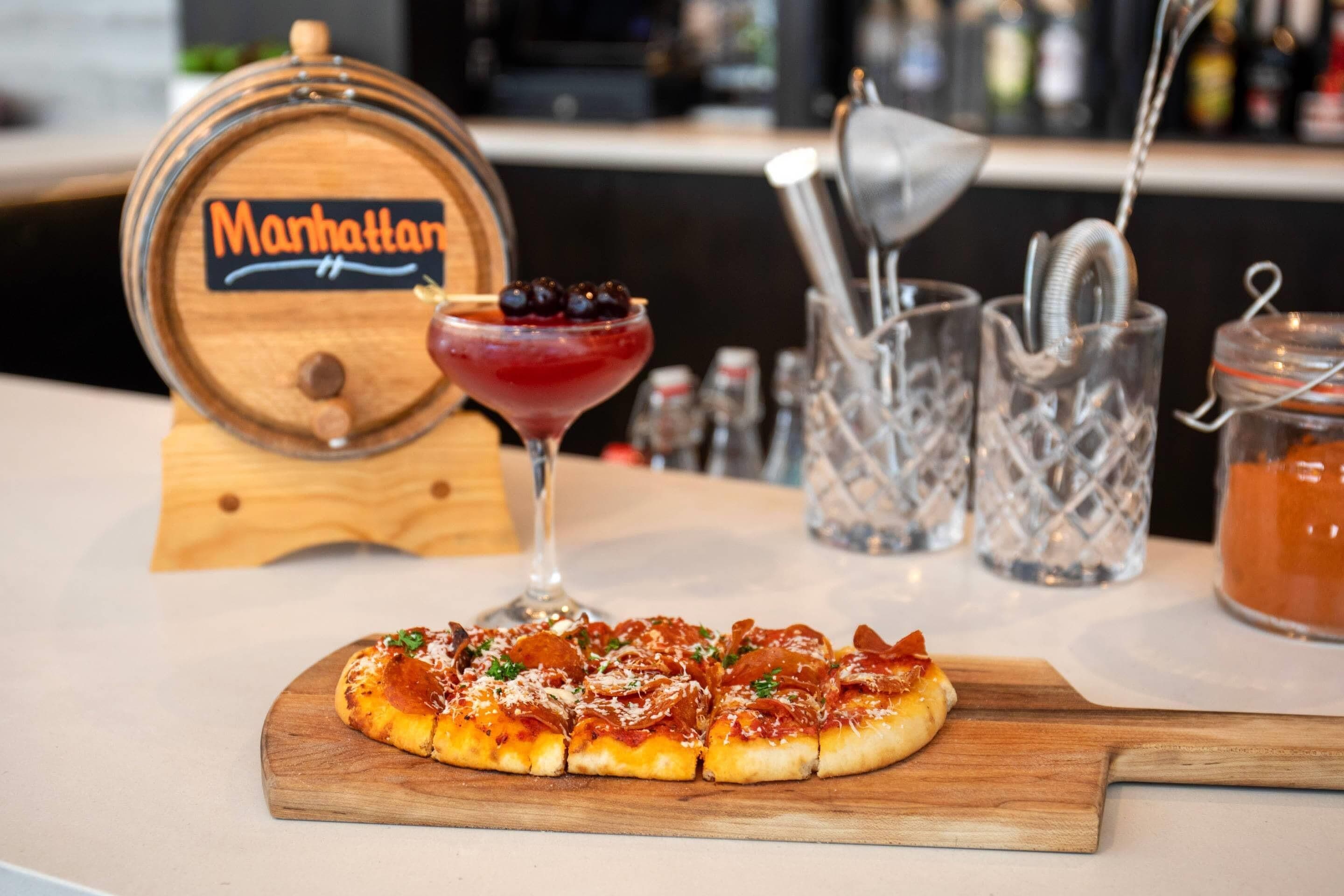 A pizza on a wooden board with a Manhattan cocktail in the background.