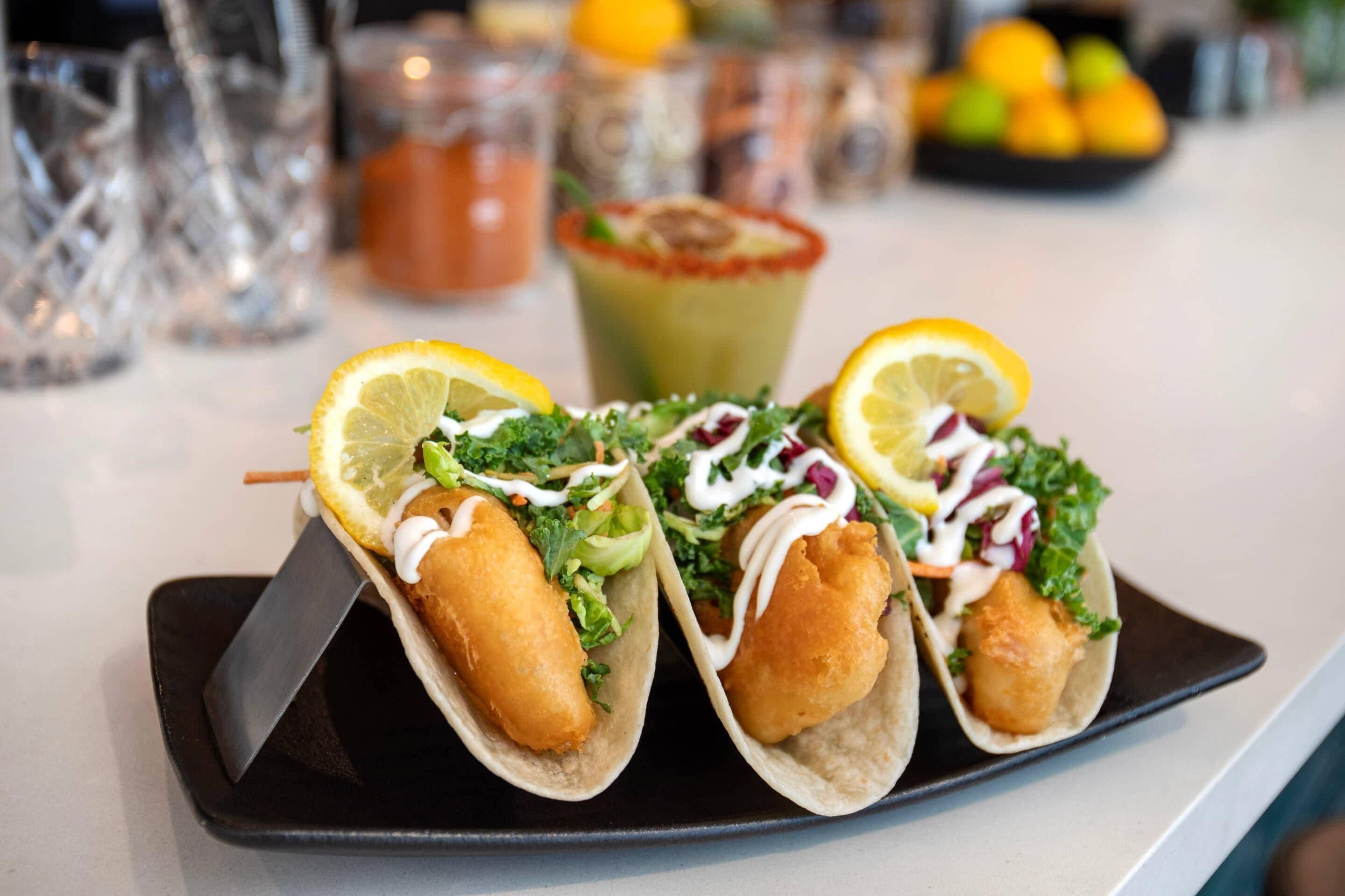 Trois tacos aux fruits de mer, servis sur une assiette noire, accompagnés d’une sauce verte et d’un quartier de citron.