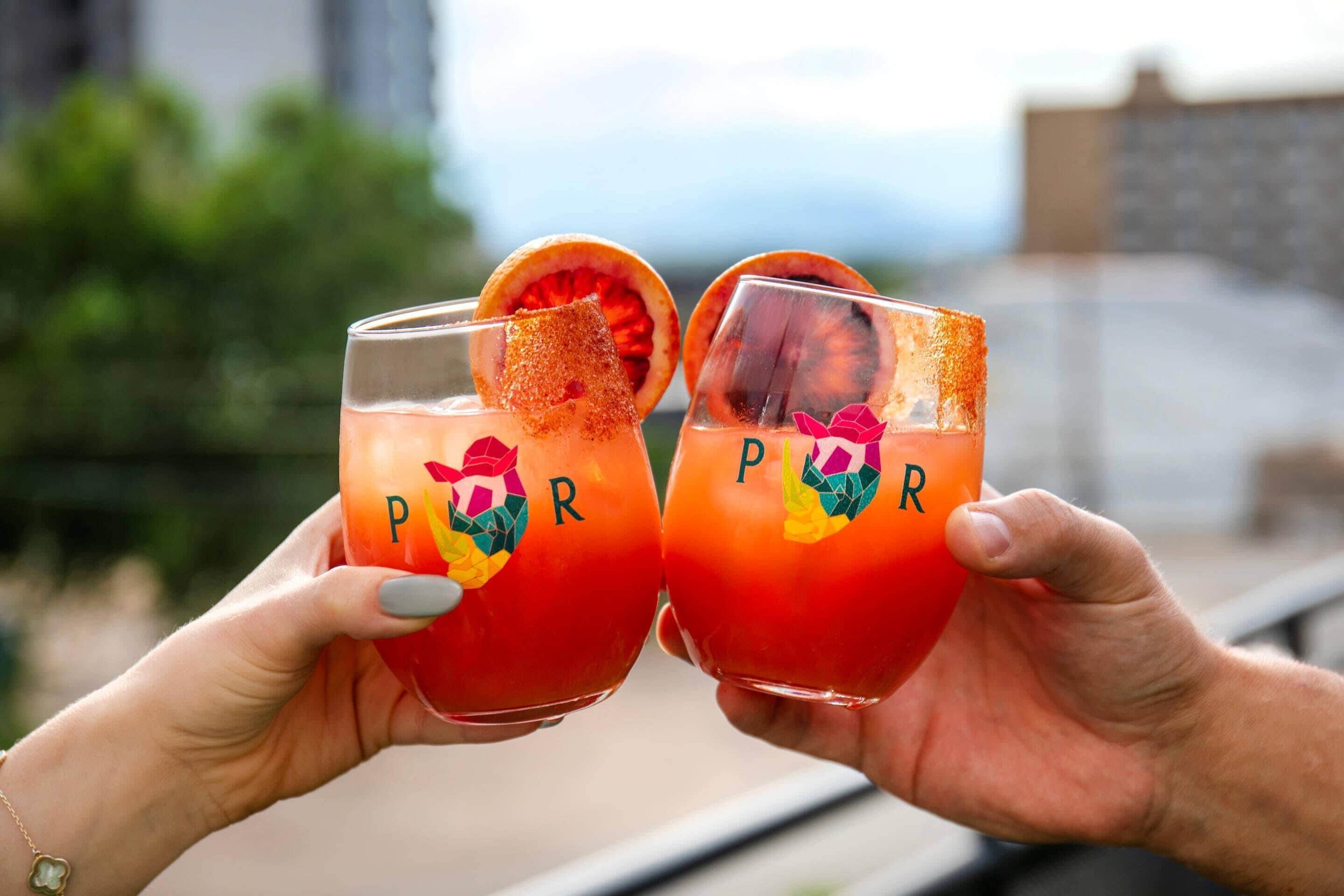 Two hands holding glasses with a tropical drink and a decorative fruit slice.