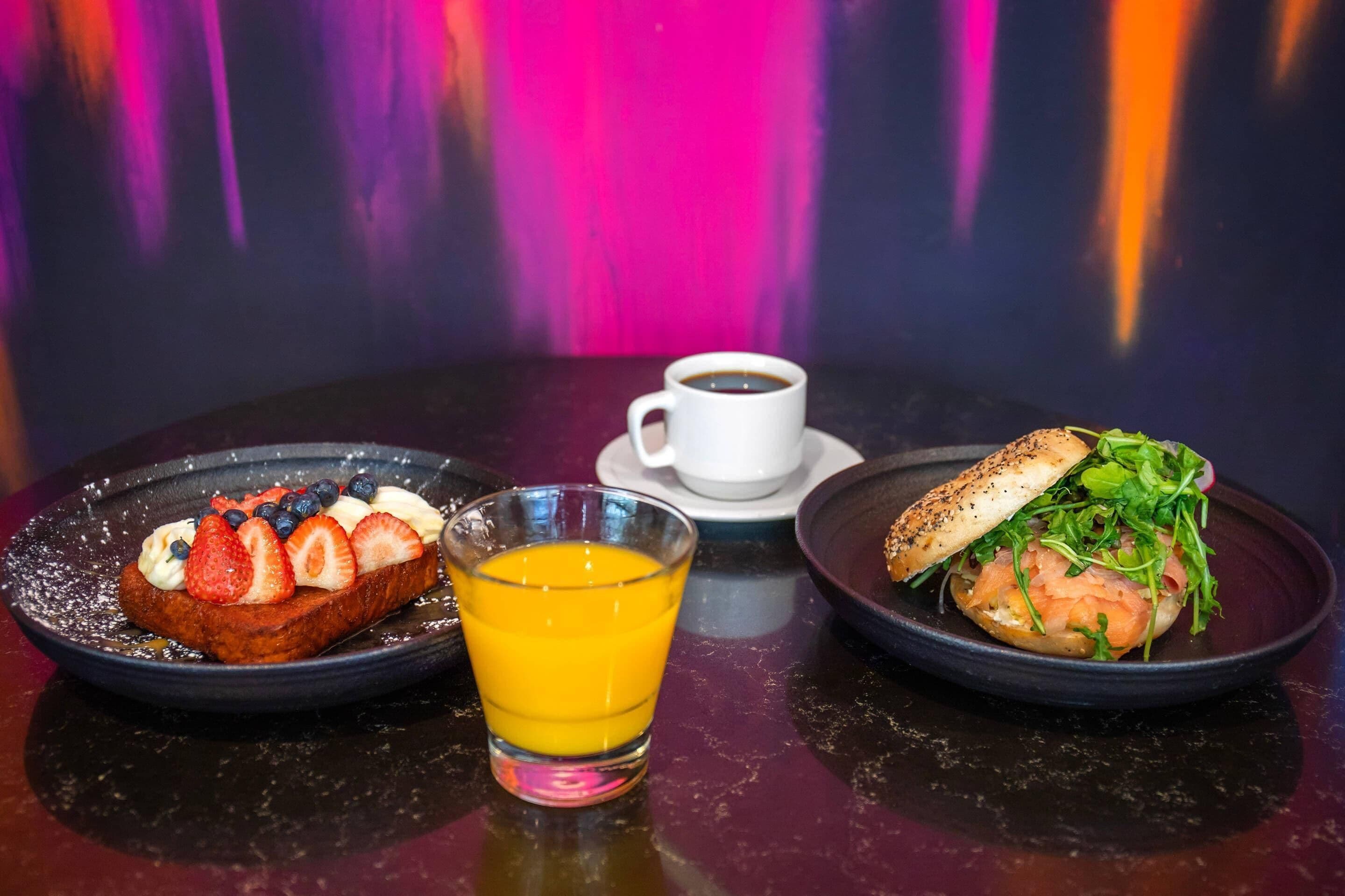 A breakfast setting with toast topped with strawberries and blueberries, a glass of orange juice, and a sandwich with greens and salmon.