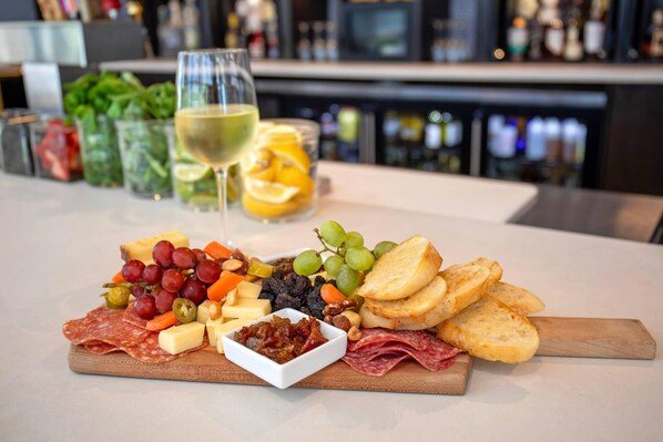 A charcuterie board with various cheeses, meats, and fruits on a wooden board, accompanied by a glass of white wine and a jar of orange slices. - Cambria Hotel Denver Downtown RiNo (Denver)