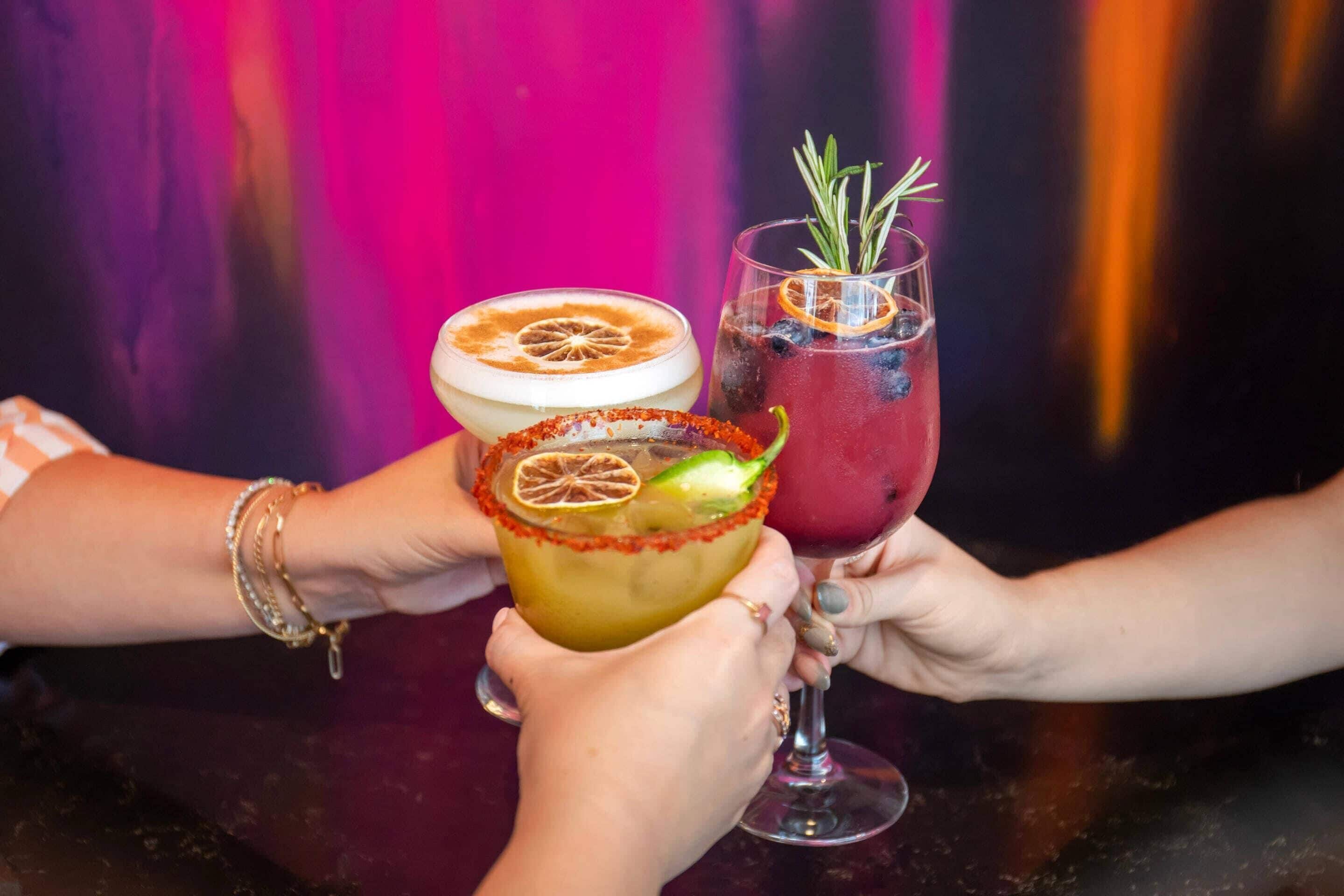 Three cocktails with garnishes, held by hands against a colorful background.