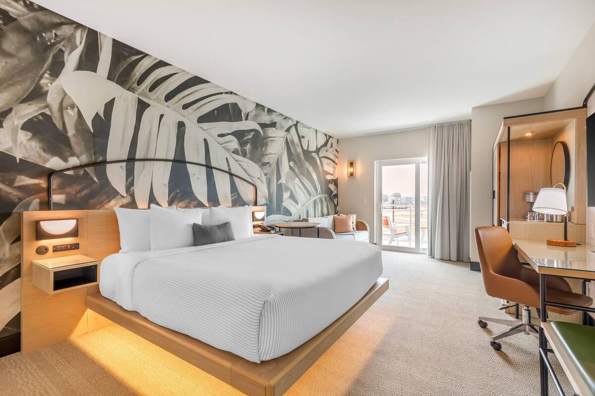 A hotel room with a large bed, a desk, a chair, a desk lamp, and a large mirror.