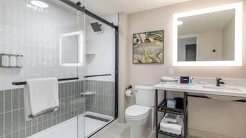 A modern bathroom with a glass-enclosed shower, a toilet, a sink with a mirror, and a shelf with towels.