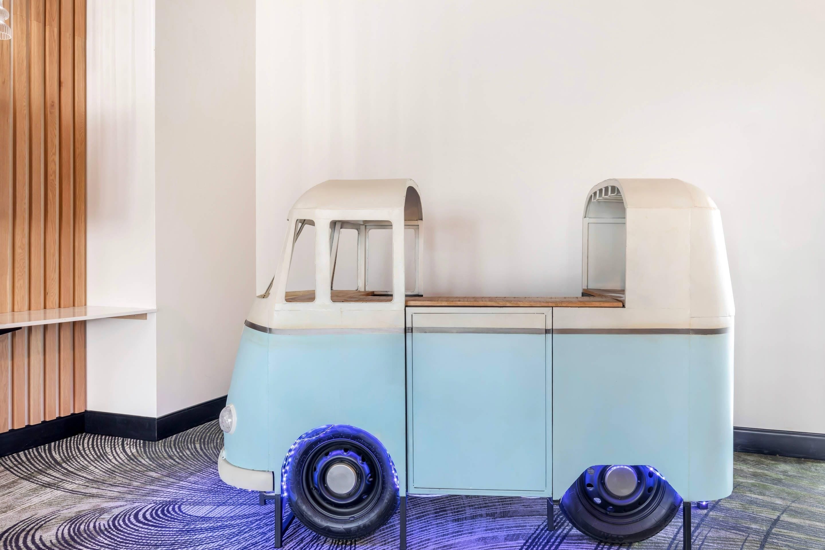 A vintage-style vehicle with a light blue and white color scheme, positioned in a room with a patterned carpet and a wooden panel wall.