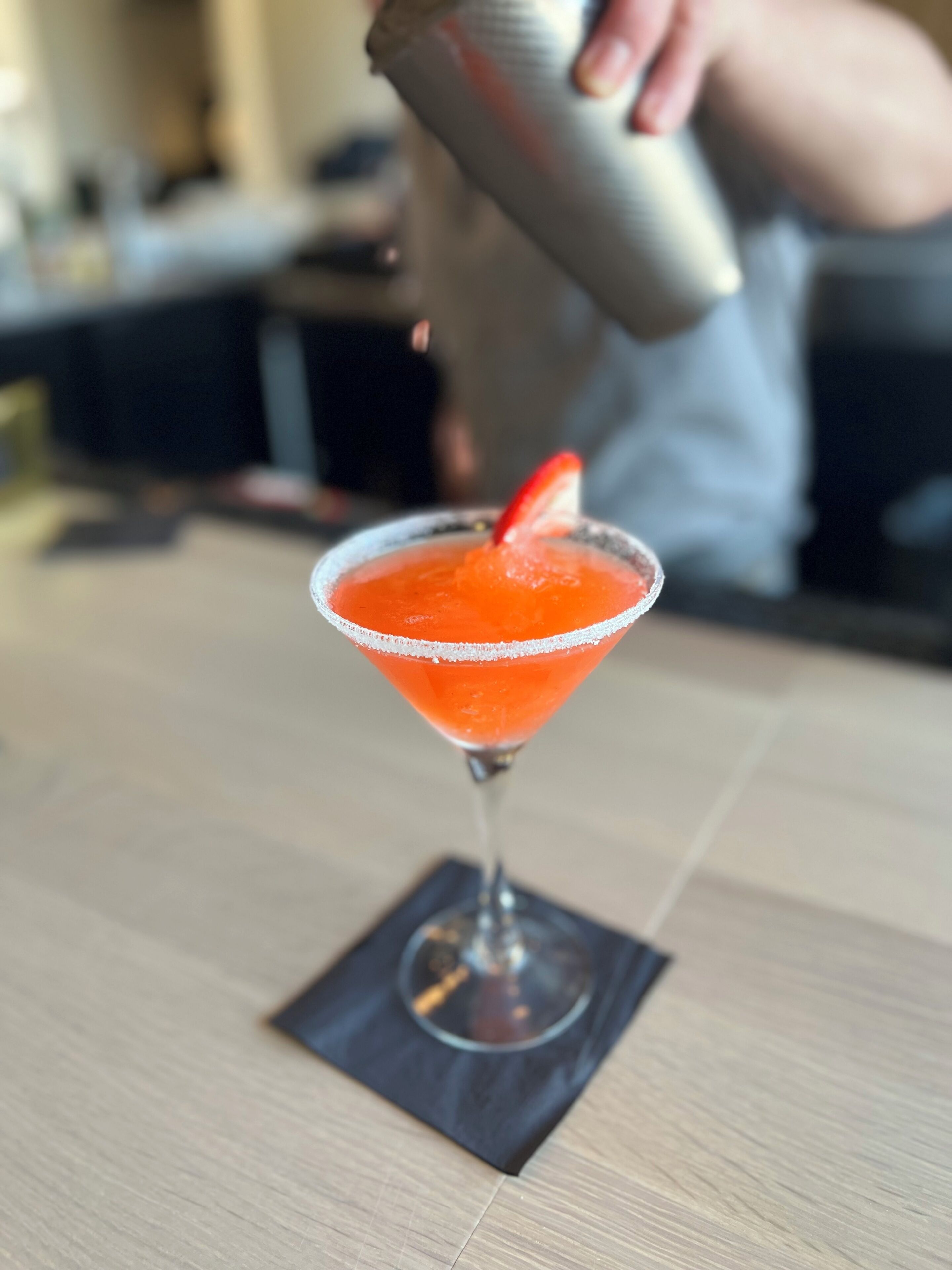 Un cóctel en una copa de martini con borde de sal y una fresa como adorno, sobre una mesa de madera.