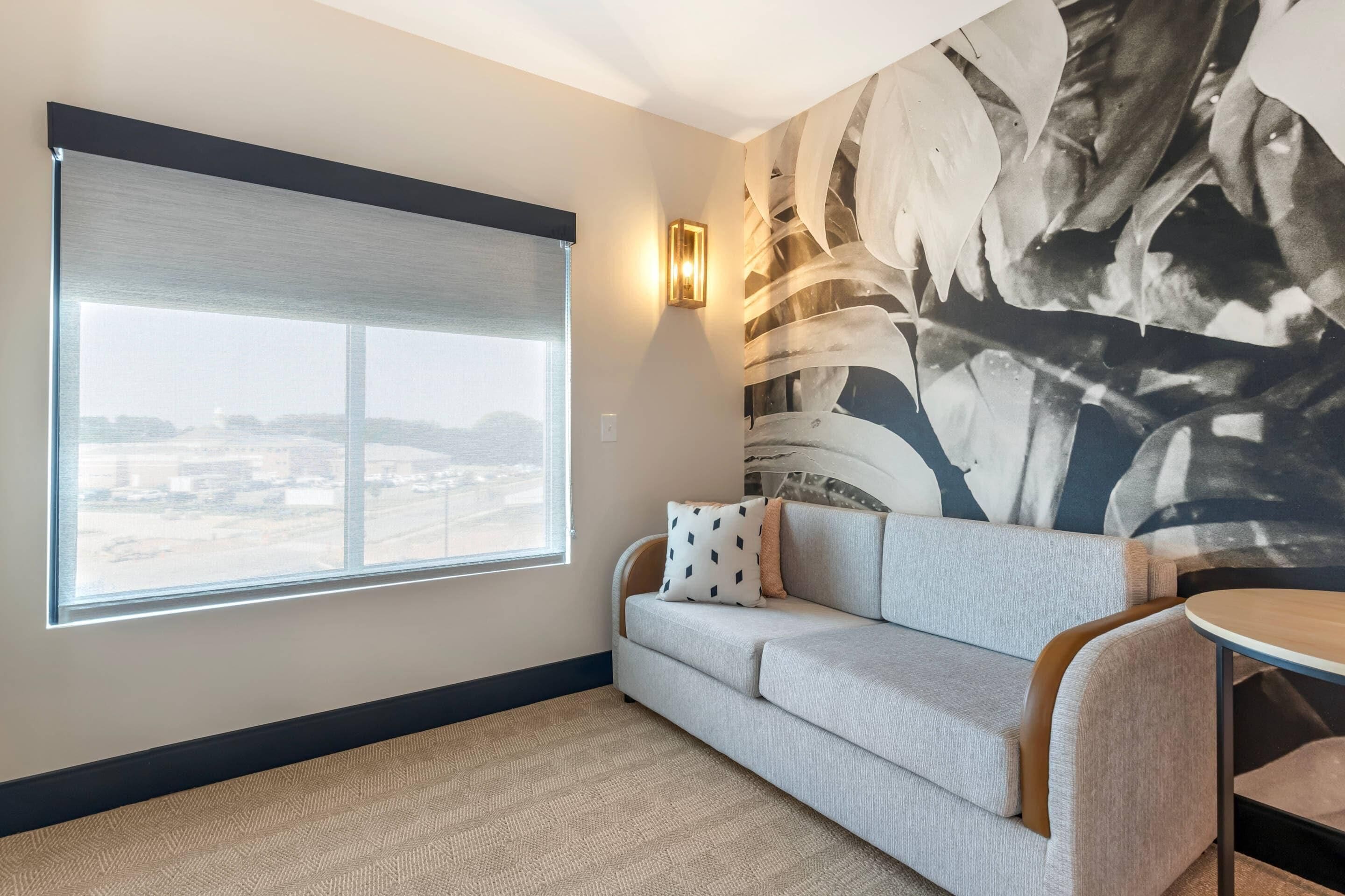 Et moderne hotelværelse med et stort vindue, en grå sofa med mønstrede puder, et rundt træbord og en væg med abstrakt kunst.