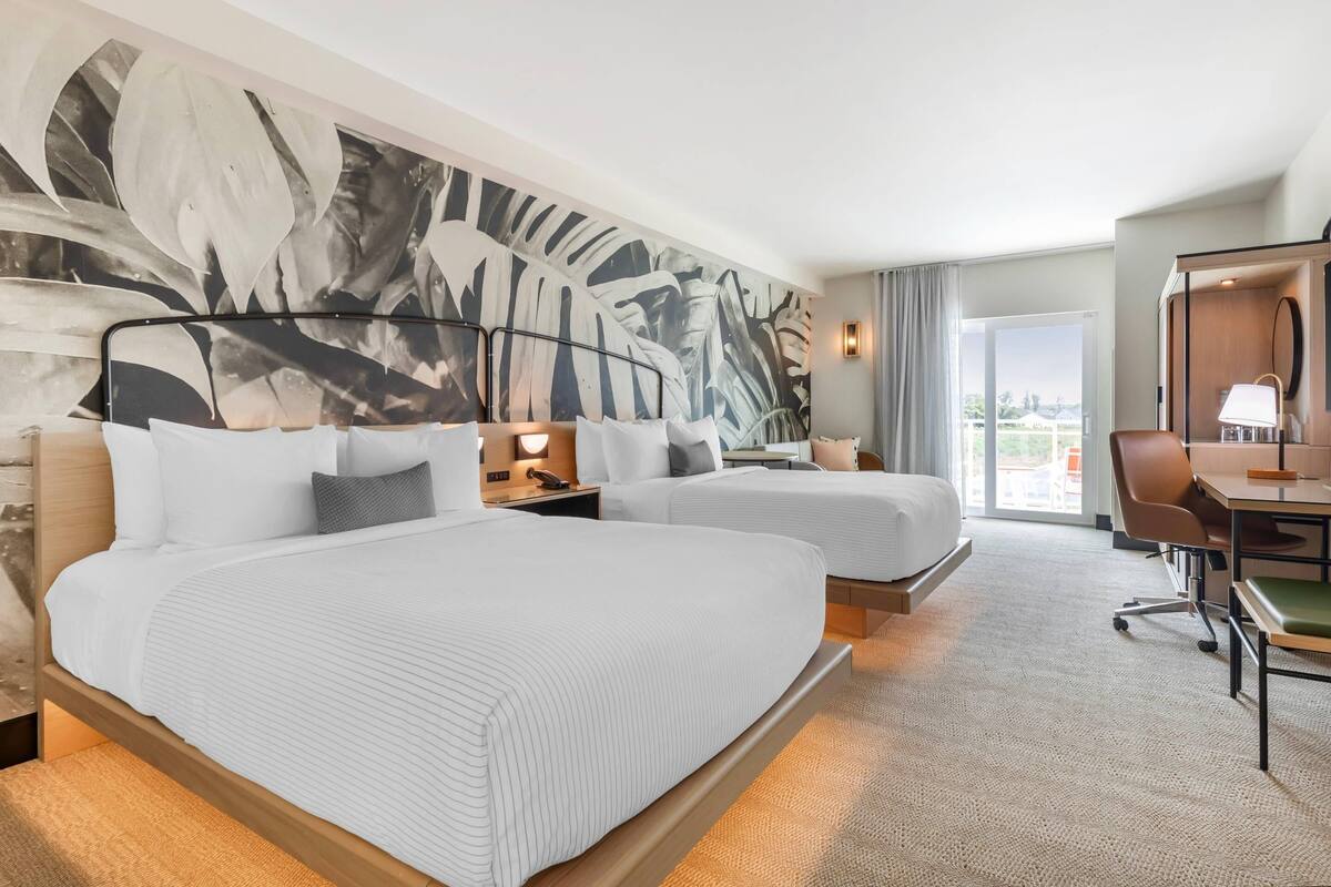 A hotel room with two beds, a desk, a chair, a large window, and a wall mural.