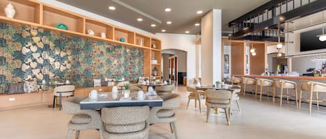 A spacious dining area with round tables and wicker chairs, a decorative wall with a leaf pattern, and a bar area with a dark staircase.