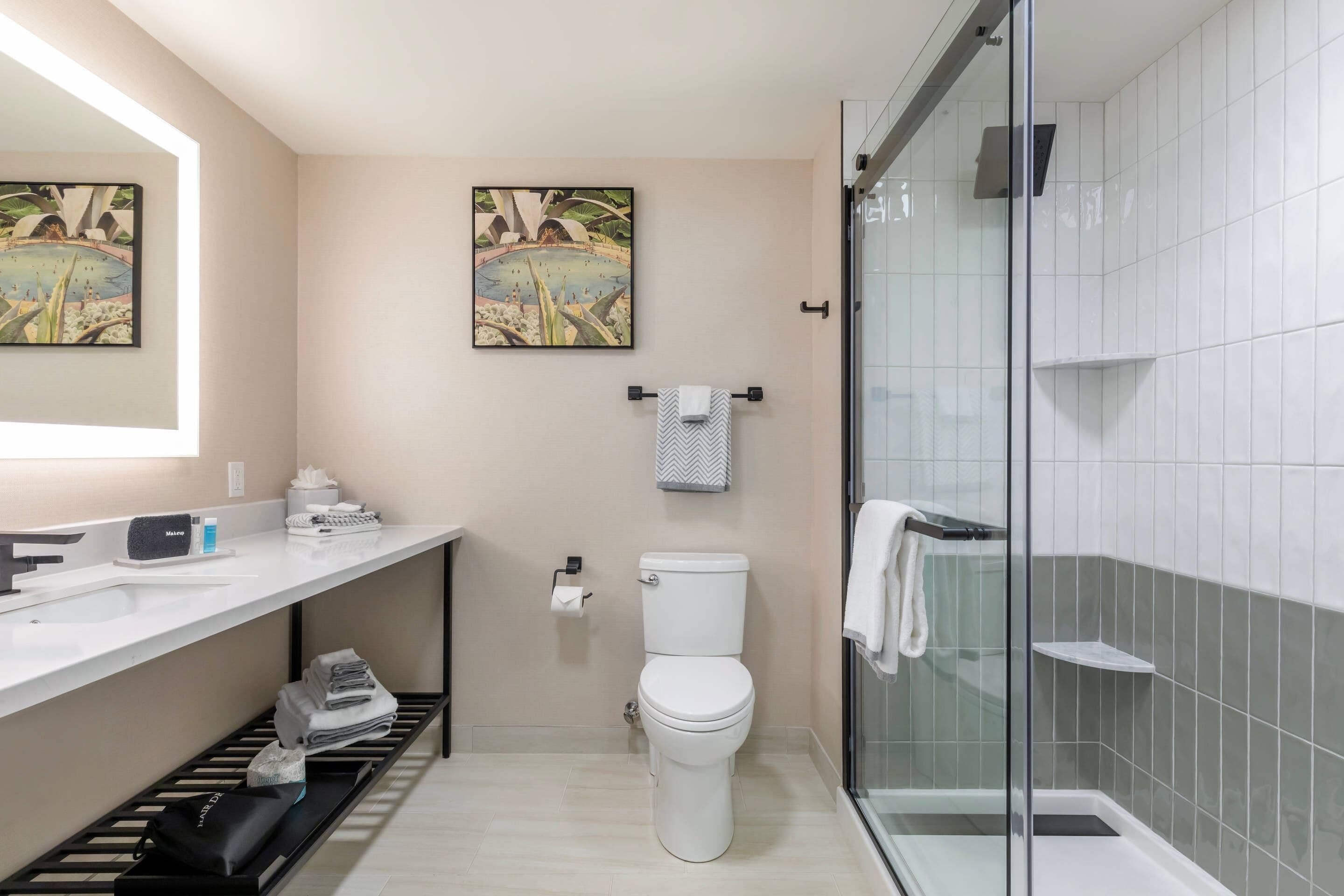 A modern bathroom with a toilet, sink, and shower. There are two framed pictures on the wall, a towel rack, and a shelf with toiletries.