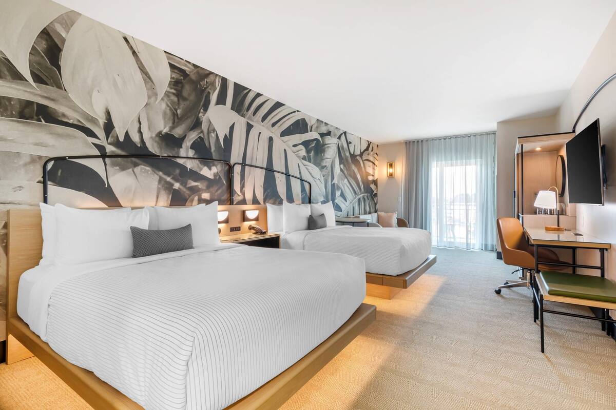 A hotel room with two beds, a desk, a chair, a TV, and a large abstract wall mural.