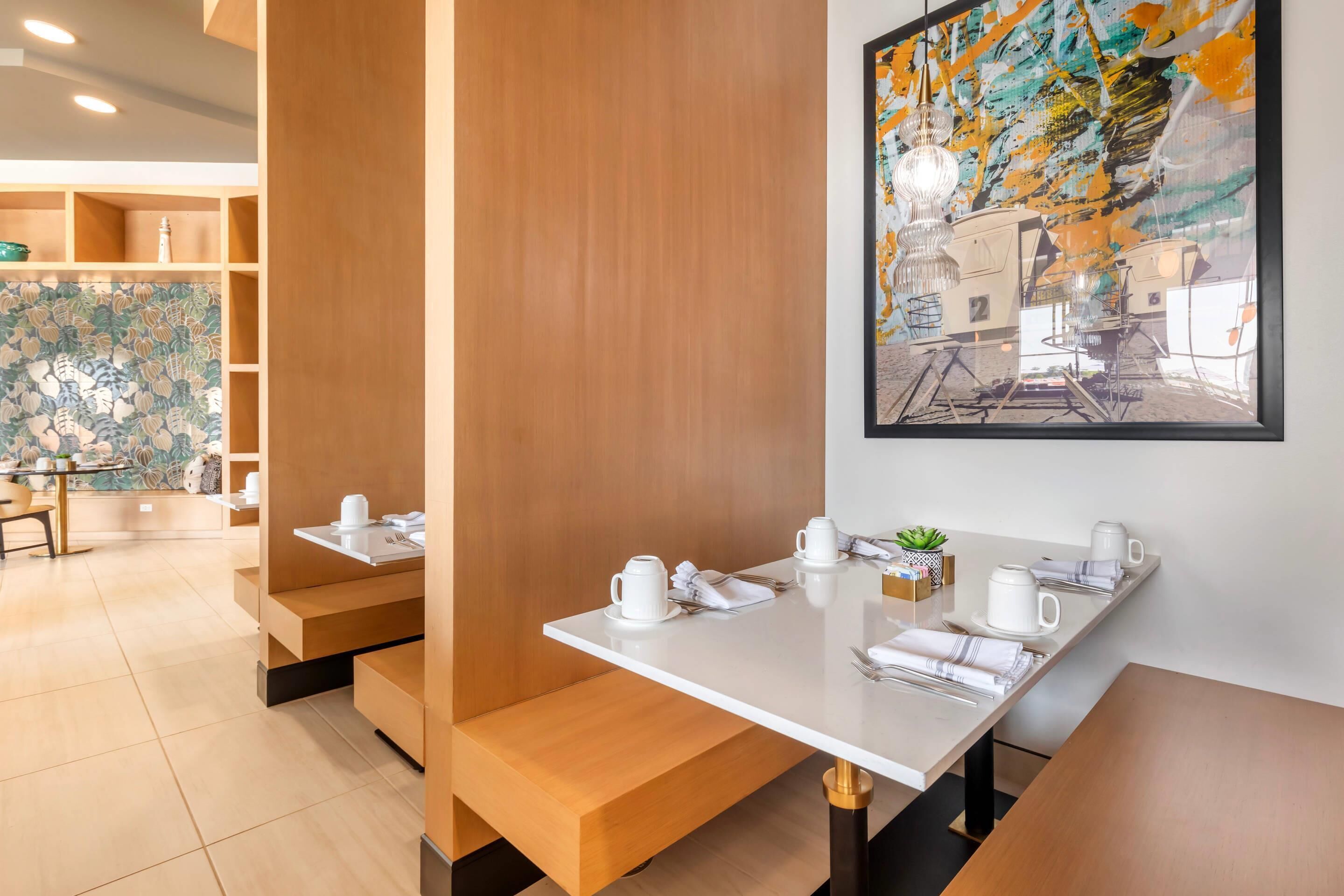 Uma sala de jantar moderna com bancos e mesas de madeira, uma grande pintura abstrata na parede e vista para a cidade através da janela.