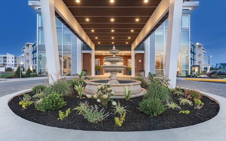 A modern building with a central fountain, surrounded by landscaped garden beds and a covered walkway.
