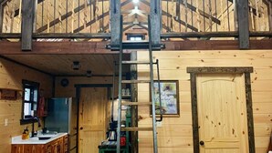A rustic cabin interior with a lofted ceiling, wooden floors, a kitchen area, and a ladder leading to a loft space. - Briar Patch Cabin RRG: Firepit, Sunset, WIFI (Stanton)