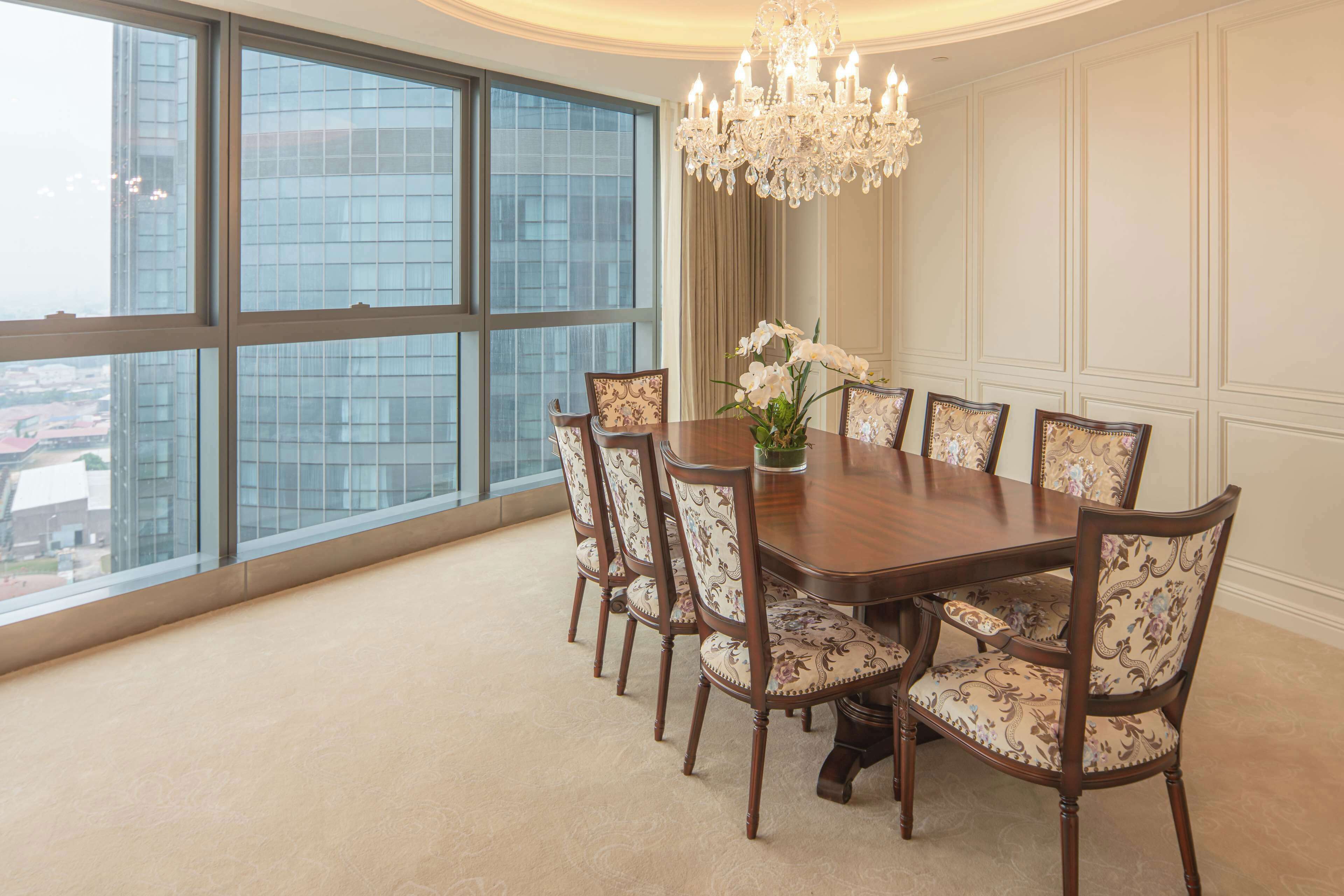 A dining room with a large table, six chairs, a chandelier, and a view of a cityscape through large windows.